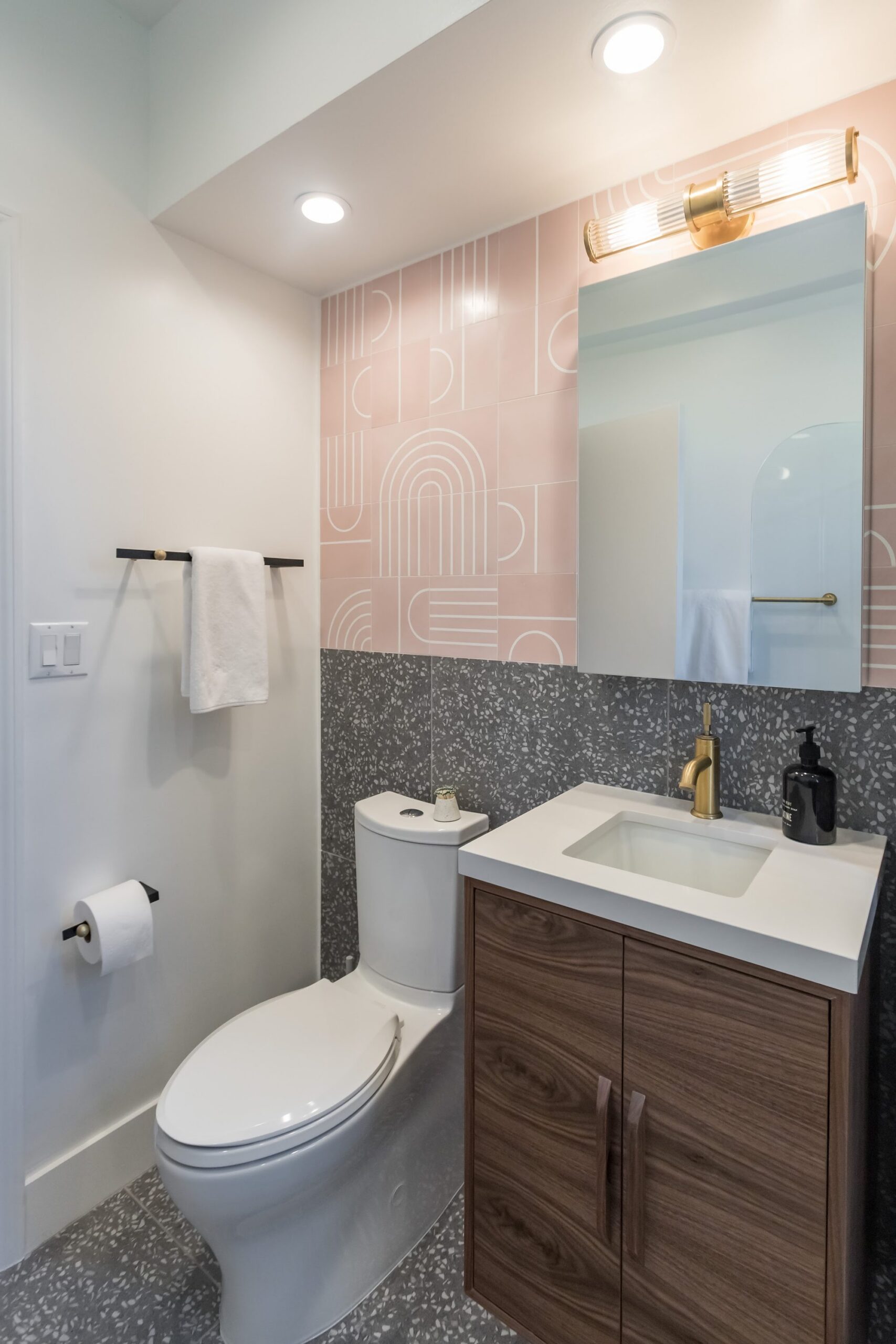 Remodeled bathroom with patterned pink tile accent wall, terrazzo-look tile, walnut vanity, and brass fixtures