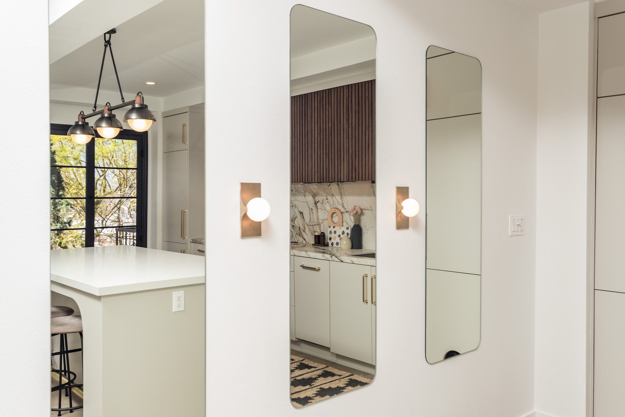 Backlit wall mirrors and brass wall sconces in a hallway opening onto a remodeled kitchen with a waterfall marble backsplash